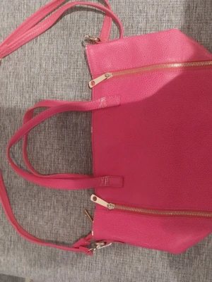Rosa handväska med gulddetaljer - Snygg rosa handväska i konstläder med två handtag och en avtagbar axelrem. Väskan har guldfärgade dragkedjor och detaljer, samt en strukturerad yta. Perfekt för dig som vill ha en färgklick till din outfit.