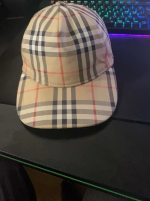 Beige rutmönstrad keps - Snygg keps i beige med klassiskt rutmönster i svart, vitt och rött. Kepsen har böjd skärm och justerbar baksida. Tillverkad i ett slätt tyg som ger en stilren look. Perfekt accessoar för att lyfta din outfit.