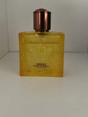Versace Eros Energy Eau de Parfum - Versace Eros Energy Eau de Parfum i en fyrkantig, gul glasflaska med elegant mönster och Medusa-huvud på framsidan. Flaskan har en metallisk, kopparfärgad kork som ger en lyxig känsla. Perfekt för dig som gillar exklusiva dofter och snygg design på parfymflaskan.
