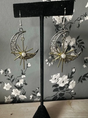 Måne och stjärna örhängen med pärla - Unika örhängen i silver- och guldfärgad metall med halvmåneform och dekorativa stjärnor. I mitten sitter en vit pärla som ger en extra touch. Perfekt för dig som gillar statement-smycken med magisk vibe. Krokar i metall för enkel användning.