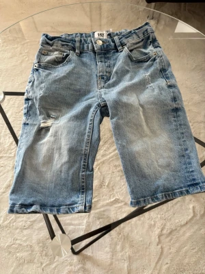 Blå jeansshorts från 157 - Säljer ett par klassiska blå jeansshorts från 157 med fem fickor och slitna detaljer på ena benet. Shortsen har normal passform och är tillverkade i bomull. Perfekta för varma dagar och enkel att matcha med olika stilar.
