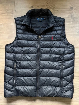 Svart dunväst från Polo Ralph Lauren - Snygg svart dunväst från Polo Ralph Lauren med hög krage och två dragkedjeförsedda fickor. Västen har ett quiltat mönster och den klassiska röda loggan broderad på bröstet. Perfekt för lager-på-lager och håller dig varm under kyliga dagar.