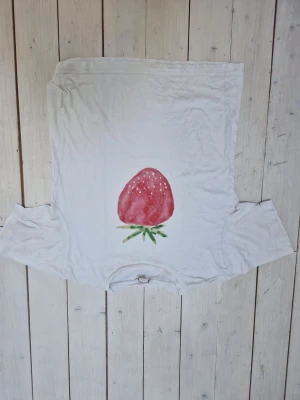 Vit t-shirt med jordgubbstryck från Monki - Säljer en vit t-shirt från Monki i bomull med ett stort jordgubbstryck framtill. Klassisk passform och korta ärmar. Perfekt för dig som gillar söta och lekfulla motiv på dina kläder. Har några minimala fläckar men syns knappt! 
