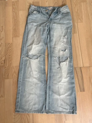 Ljusblå slitna wide leg jeans - Säljer ett par ljusblå jeans med breda ben och flera slitningar och hål på framsidan. Jeansen har klassisk femficksmodell och en avslappnad, trendig passform. Perfekta för en cool och avslappnad stil.