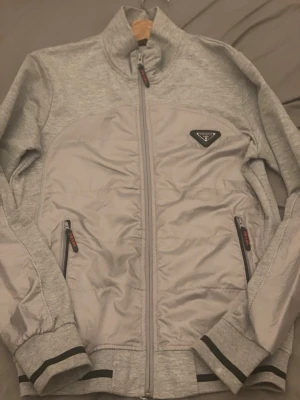 Prada kofta  - Snygg grå jacka från Prada med quiltad front och två fickor med dragkedja. Jackan har hög krage, långa ärmar och svarta detaljer på muddar och dragkedjor. Perfekt för dig som vill ha en stilren och exklusiv look.