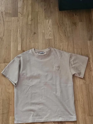 Beige t-shirt från Dior - Snygg beige t-shirt från Dior med diskret broderad logga på bröstet. Klassisk rund halsringning och korta ärmar. Tillverkad i mjuk bomull för en skön känsla och clean look.