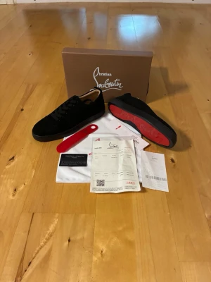 Svarta sneakers från Christian Louboutin - Snygga svarta sneakers från Christian Louboutin med klassisk röd sula och diskret logga. Skorna har en låg profil, svart mocka och svarta skosnören. Perfekta för dig som vill ha en stilren och exklusiv look.