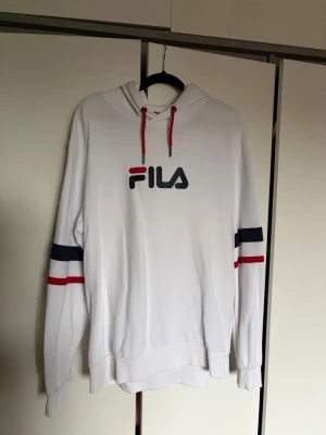 Vit hoodie från FILA med logga - Snygg vit hoodie från FILA med stor logga på bröstet och färgdetaljer i rött och marinblått på ärmarna. 