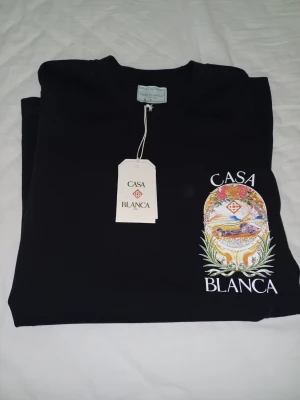 Svart Casa Blanca t-shirt med tryck - Snygg svart t-shirt från Casa Blanca med färgglatt tryck framtill som visar en bil, blommor och logga. T-shirten har rund hals och korta ärmar. Perfekt för dig som vill sticka ut med en unik design.