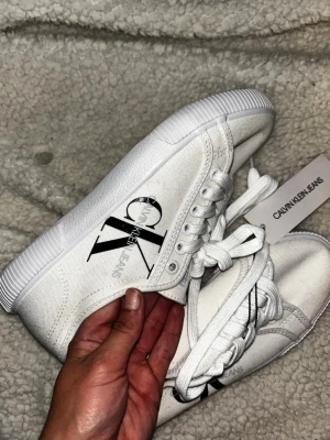 Vita Calvin Klein Jeans sneakers - Snygga vita sneakers från Calvin Klein Jeans med svart CK-logga på sidan. Skorna har klassisk låg profil, vita snören och sula i gummi. Ovandelen är i textil vilket ger en clean och fräsch look. Perfekta för dig som gillar stilrena och trendiga skor.
