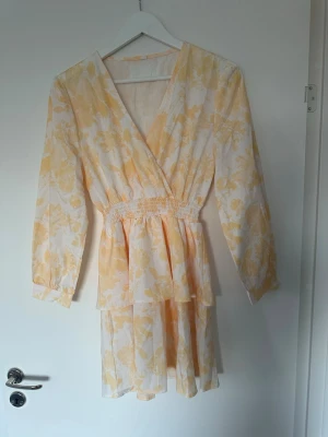 Kort blommig omlottklänning - Söt kort klänning med blommigt mönster i ljusgult och vitt. Klänningen har omlottmodell, v-ringning, långa ärmar och smock i midjan för en bekväm passform. Perfekt för varma dagar och somriga tillfällen.