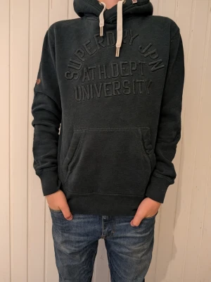 Mörkgrå hoodie med broderad text - Mörkgrå hoodie från Superdry med broderad text 'Superdry JPN ATH. DEPT UNIVERSITY' på bröstet. Klassisk känguruficka framtill, ribbade muddar och vit dragsko i huvan. Perfekt för en avslappnad och cool stil.