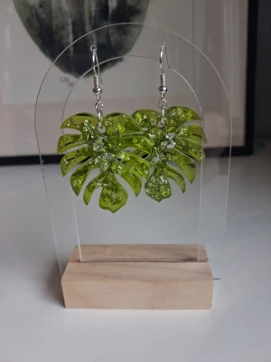 Gröna monstera örhängen i resin/epoxy handgjorda - Unika örhängen i form av monstera-blad, gjorda i transparent grön resin med inslag av silverfolie detaljer. Öronkrokenärnickelsäker. Perfekta för dig som vill sticka ut med ett tropiskt och trendigt smycke. Längd ca 4cm långa