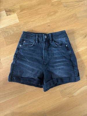 Svarta högmidjade jeansshorts - Snygga svarta högmidjade jeansshorts med fem fickor och uppvikta benslut. Klassisk knapp- och dragkedjestängning framtill. Perfekta för varma dagar och enkel att matcha med olika stilar.
