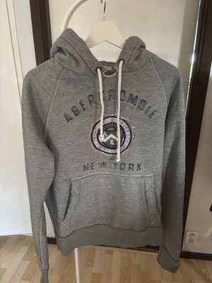 Archive Grå hoodie Abercrombie & Fitch - Snygg grå hoodie från Abercrombie & Fitch med broderad text 'Abercrombie New York' och ett emblem på bröstet. Tröjan har huva med vita snören och en stor magficka. Tillverkad i mjukt sweatshirtmaterial, perfekt för chill dagar.