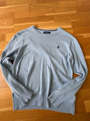 Ljusblå stickad tröja från Polo Ralph Lauren - Ljusblå stickad tröja från Polo Ralph Lauren med rund halsringning och diskret broderad logga på bröstet. Tröjan har långa ärmar och ribbade muddar vid ärmslut och nederkant. Perfekt för lager på lager och enkel att matcha.