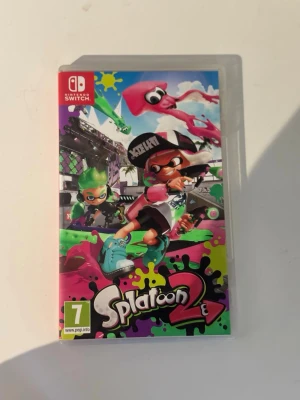 Splatoon 2 till Nintendo Switch - Splatoon 2 till Nintendo Switch är ett färgstarkt och actionfyllt spel där du tävlar i lagbaserade bläckstrider. Omslaget har neonfärger med grönt, rosa och svart och spelet levereras i original plastfodral med kassett.