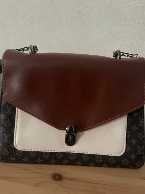 Luise Vuitton Brun och vit axelväska med kedja - Snygg axelväska i brunt, vitt och mörkbrunt med klassiskt mönster. Väskan har ett lock i brunt läder, vit frontficka och kedjerem. Perfekt storlek för det viktigaste och stängs med ett metallspänne. Insidan har ett större fack och en mindre ficka.