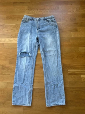 Ljusblå raka jeans från Friday Denim - Ljusblå jeans med rak passform från Friday Denim. Jeansen har en sliten look med ett stort hål på vänster lår och klassiska fem fickor. Tillverkade i denim med synliga sömmar och knappgylf. Perfekta för en avslappnad och trendig stil.