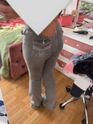 Grå bootcut jeans med detaljer - Säljer ett par grå bootcut jeans med snygga broderade detaljer och knappar på bakfickorna. Jeansen har hög midja och utsvängda ben, vilket ger en retro vibe. Perfekta för dig som vill sticka ut lite extra med coola detaljer.