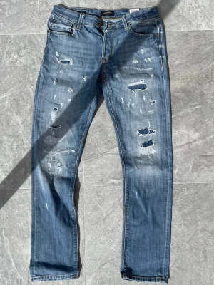 Jeans med slitningar  - Riktigt snygga Jack and Jones jeans med slitningar. Storlek, mått och modell syns på bilderna. Ej slitet i skrevet. Skickar snabbt! 