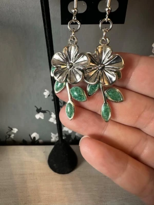 Silvriga blomörhängen med gröna blad - Unika örhängen i form av blommor med silvrig metall och detaljerade gröna blad. Blommorna har en tydlig struktur och hänger snyggt från örat. Perfekta för dig som vill ha ett statement-smycke med naturinspirerad design.