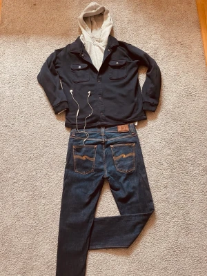 Mörkblå Nudie Jeans - Säljer ett par feta Nudie Jeans i klassisk rak modell med snygga kontrastsömmar och vågigt broderi på bakfickorna. Jeansen har fem fickor och bälteshällor. Materialet är slitstark denim i bomull. Perfekta till en schysst laidback look. Storlek = W31, L32. 
