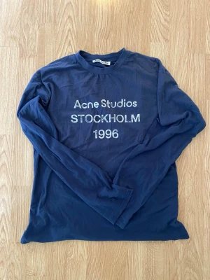 Mörkblå långärmad topp Acne Studios - Mörkblå långärmad topp från Acne Studios med tryck 'Acne Studios STOCKHOLM 1996' i vitt framtill. Klassisk rund halsringning och avslappnad passform. Perfekt för dig som gillar stilrena och trendiga plagg.