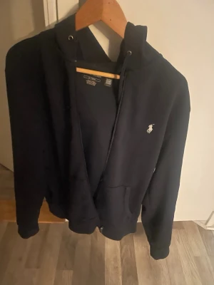 Ralph lauren kofta  - Säljer en svart hoodie med dragkedja och huva. Klassisk design med fickor framtill och en liten broderad logga på bröstet. Perfekt för chill dagar eller när du vill ha en enkel och stilren look. Mjuk bomullskänsla och ribbade muddar vid ärmar och midja.