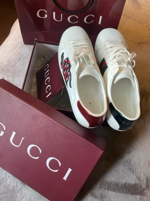 Gucci Aces - Vita Gucci Ace sneakers i läder med klassiska gröna och röda sidoremsor och broderad orm på utsidan. Baktill har ena skon röd och andra grön metallic-detalj. Snygg och ikonisk modell med vita snören och låg profil.