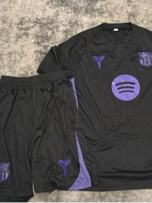 Svart och lila fotbollsställ - Säljer ett svart fotbollsställ med lila detaljer, bestående av tröja och shorts. Setet har tryckta loggor och mönster i lila, inklusive klubbmärke och sponsortryck. Materialet är lätt och andas, perfekt för träning eller match.