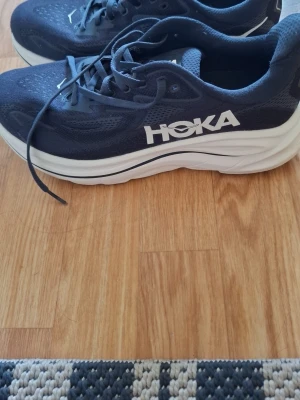 Mörkblå Hoka sneakers med tjock sula - Snygga mörkblå sneakers från Hoka med vit, tjock sula och ventilerande mesh-ovandel. Skorna har klassisk snörning och stor Hoka-logga på sidan. Perfekta för dig som gillar sportig stil och komfort.