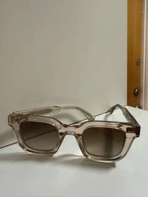 Chimi glasögon  - Snygga solglasögon med transparent beige båge och fyrkantig form. Glasen är lätt tonade i brunt och ger en cool retrovibe. Perfekta för soliga dagar när du vill sticka ut lite extra.