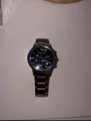 Emporio Armani silverfärgad klocka - Stilren klocka från Emporio Armani med silverfärgad länk i metall och svart urtavla. Klockan har tre mindre urtavlor, datumvisning och tydliga markeringar. Perfekt accessoar för dig som gillar en klassisk men modern look. Ar2448