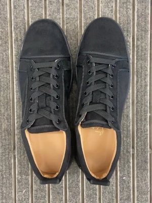 Svarta Louboutin Skor - Snygga svarta sneakers från Louboutin med klassisk låg profil och ikonisk röd sula. Skorna har svarta skosnören, ovandel i mocka och läderdetaljer samt beige innersida med louboutin texten. Perfekt för dig som vill sticka ut med exklusiva skor. Skorna är använda men däremot i mycket bra skick, med gott om liv kvar. Då menar jag att skorna på bilden är när dem var nya och nu har dem blivit använda ett par gånger och ser inte lika nya ut.