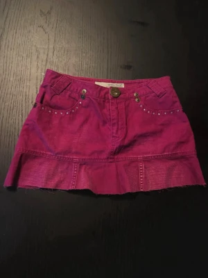 Rosa jeanskjol med volang  - Vet ej vad strl 14 är men skulle säga XS! Snygg rosa kort jeanskjol med dekorativa stenar vid fickorna fram och dekorativa sömmar. Kjolen har två bakfickor med strassdetaljer och rå kant nedtill för en trendig look. Perfekt för dig som vill sticka ut lite extra. 