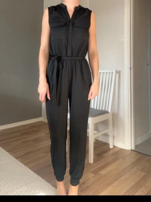 Svart ärmlös jumpsuit med knytskärp - Snygg svart ärmlös jumpsuit med två bröstfickor och knytskärp i midjan. Jumpsuiten har en avslappnad passform, smala ben och ståkrage. Perfekt för dig som gillar stilrena och bekväma plagg. Stl 32/34 men passar även stl 36 beroende hur man vill att den ska sitta. 