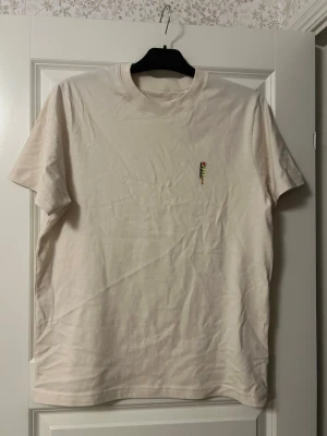 Beige t-shirt med Twister-glass tryck - Säljer en beige t-shirt i mjuk bomull med ett litet Twister-glass tryck på bröstet. T-shirten har rund hals och korta ärmar, perfekt för dig som gillar enkla men roliga detaljer. Passar till det mesta och är riktigt skön att bära.