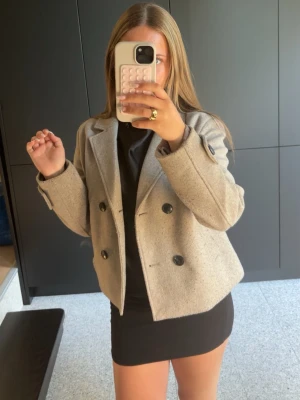 Beige dubbelknäppt kappa - Snygg beige kappa med dubbelknäppning och stora svarta knappar. Kortare modell och raka ärmar. Perfekt för dig som vill ha en stilren och trendig look till hösten.