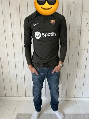 Nike FC Barcelona träningströja - Säljer en mörkgrön långärmad träningströja från Nike med FC Barcelona-logga och Spotify-tryck på bröstet. Tröjan har half zip-dragkedja och är tillverkad i ett stretchigt syntetmaterial. Perfekt för fotbollsträning eller uppvärmning.