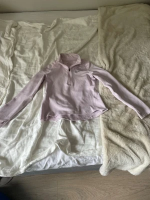 Ljuslila långärmad träningströja Nike - Ljuslila långärmad träningströja från Nike med halv dragkedja framtill och diskret logga på bröstet. Tröjan har en figurnära passform och är tillverkad i ett lätt, stretchigt material som passar perfekt för träning och sportaktiviteter.