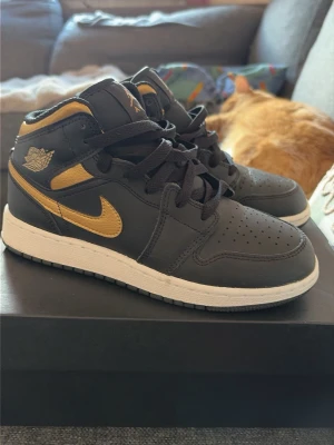 Svarta och guld Air Jordan 1 Mid - Snygga Air Jordan 1 Mid sneakers i svart läder med guldiga detaljer och vit sula. Klassisk siluett med perforerad tåbox, svarta skosnören och ikoniska loggor på sidan och plösen. Perfekta för dig som vill sticka ut med stilrena och sportiga skor.