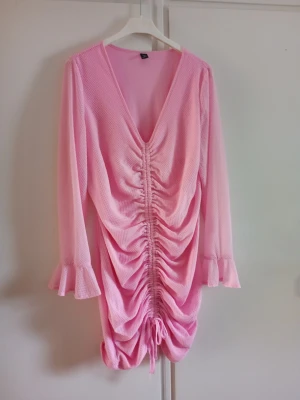 Rosa klänning med snörning drapering 4 XL - Säljer en rosa kortklänning med v-ringning och långa ärmar med volangkant. Klänningen har snygg snörning framtill som ger en rynkad effekt och en omlottkänsla. Perfekt för dig som vill sticka ut lite extra.