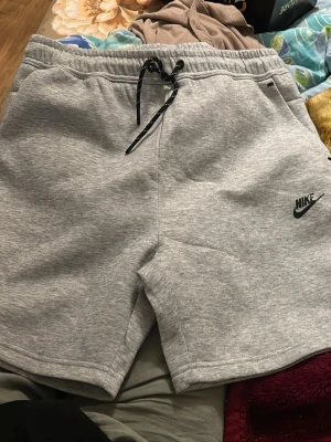 Grå sweatshorts från Nike - Säljer ett par grå sweatshorts från Nike med svart logga på vänster ben. Shortsen har snörning i midjan och är tillverkade i mjukt bomullsmaterial. Perfekta för chill dagar eller träning.