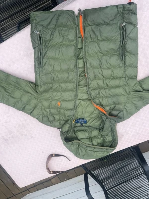 Grön dunjacka från Polo Ralph Lauren - Snygg grön dunjacka från Polo Ralph Lauren med huva och två dragkedjeförsedda bröstfickor. Jackan har orange detaljer på insidan och klassisk logga broderad på bröstet. Perfekt för kalla vinterdagar och riktigt skön passform.