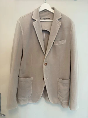 Beige kavaj från Circolo 1901 - Stilren beige kavaj från Circolo 1901 med två knappar framtill, klassisk krage och tre fickor. Tillverkad i mjukt material som ger en avslappnad men ändå dressad look. Perfekt för dig som vill ha en modern och bekväm kavaj. | Storlek: 52 (M) | Skick: 9/10 | Nypris: 6399kr | Ärmlängd: 72 cm | Axelbredd: 51 cm | Meddela vid eventuella frågor! Mvh ResellGarms 