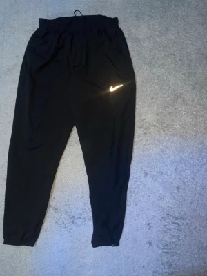 Svarta Nike Dri-FIT mjukisbyxor - Svarta mjukisbyxor från Nike med Dri-FIT-material som andas. Byxorna har resår i midjan med dragsko, muddar vid bensluten och en vit Nike-logga på benet. Perfekta för chill eller träning.