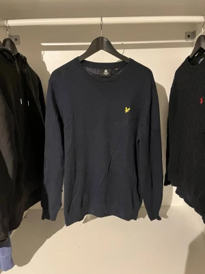 Mörkblå stickad tröja Lyle & Scott - Mörkblå stickad tröja från Lyle & Scott med rund halsringning och den klassiska gula logon broderad på bröstet. Tröjan är L men har krympt så den är storlek S. Tröjan har långa ärmar och är tillverkad i mjukt material som passar perfekt till höst och vinter. 