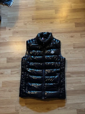 Svart glansig dunväst från Moncler - Snygg svart dunväst från Moncler med glansigt material och hög krage. Västen har en klassisk Moncler-logga på bröstet och dragkedja framtill. Perfekt för lager-på-lager och ger en trendig look till din outfit.