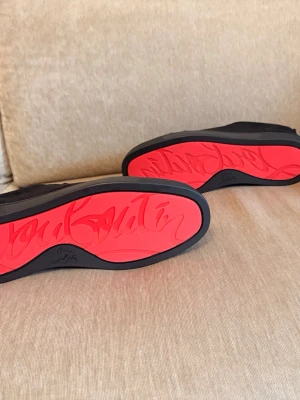 Svarta sneakers med röd sula - Snygga svarta sneakers från Christian Louboutin med ikonisk röd sula och diskret logga undertill. Skorna har en klassisk låg profil, snörning och är tillverkade i mocka med en mjuk insida. Perfekt för dig som vill sticka ut med stil.